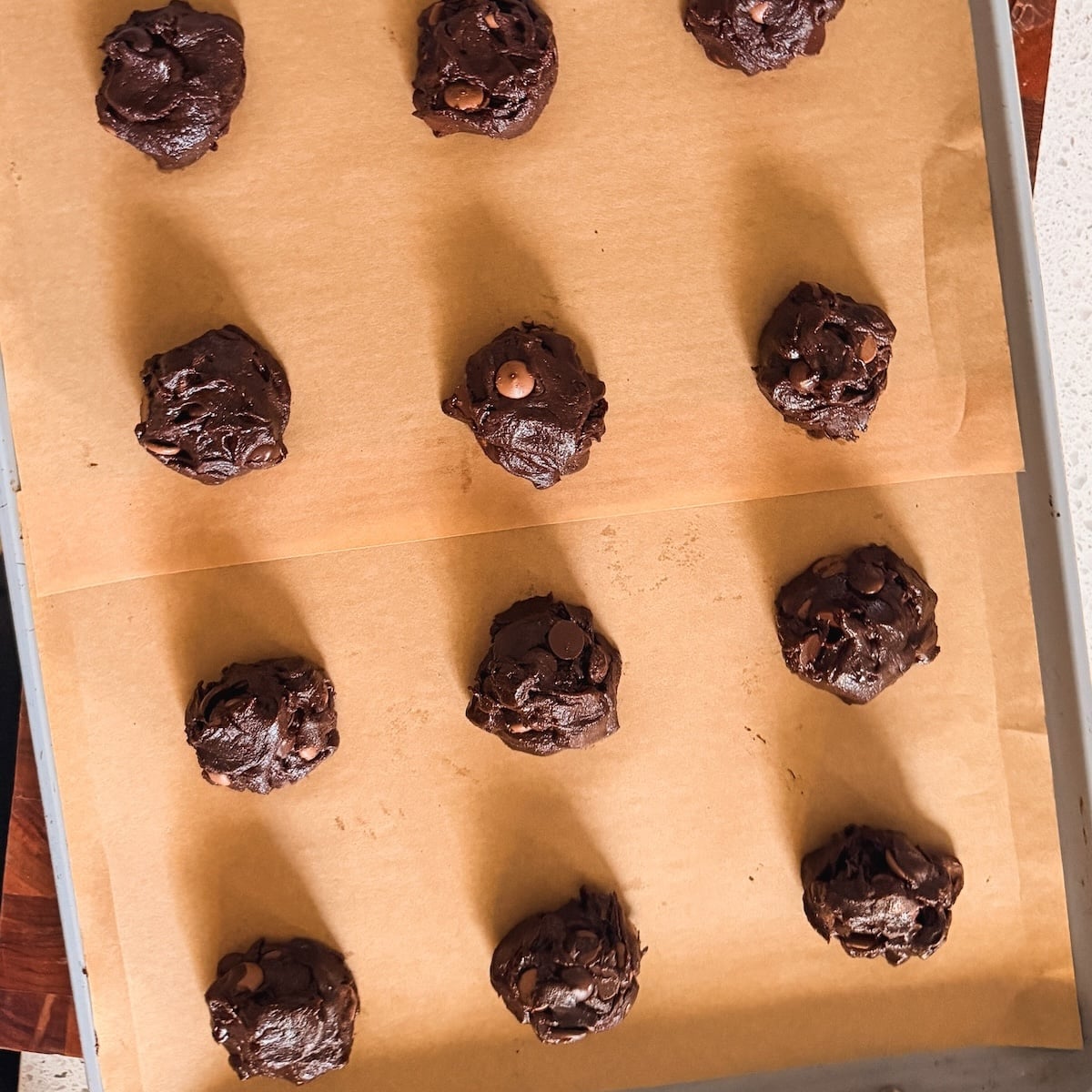 Twelve scoops of chocolate cookie dough with chocolate chips are evenly spaced on two sheets of brown parchment paper, ready to be baked on a metal tray.