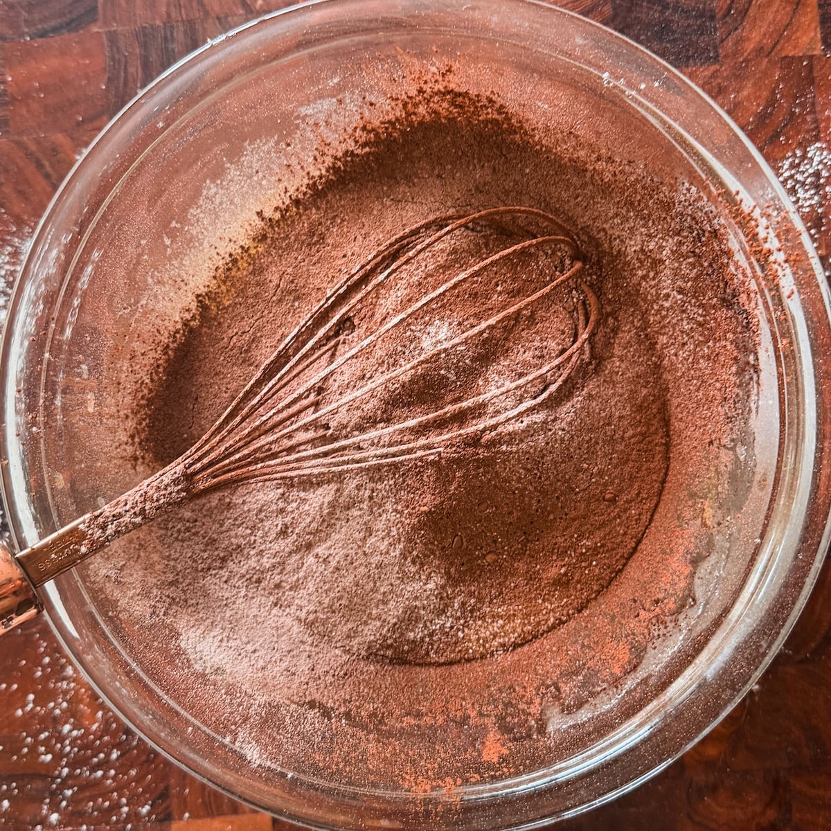 A glass bowl filled with cocoa powder and flour being mixed with a metal whisk sits on a wooden surface, with some flour scattered around the bowl.