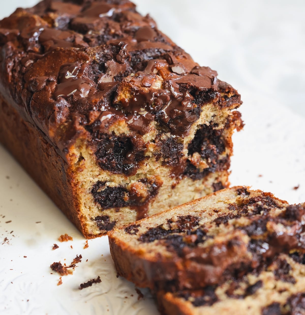 A close-up of a sliced loaf of brownie banana bread on a white surface, showing gooey melted chocolate chunks and a moist, textured interior.