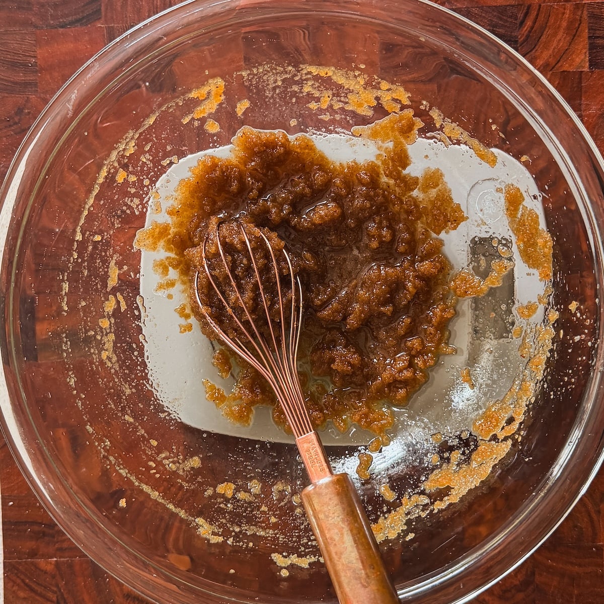 A glass mixing bowl on a wooden surface holds a partially mixed combination of brown sugar and a creamy liquid, with a copper whisk resting inside.