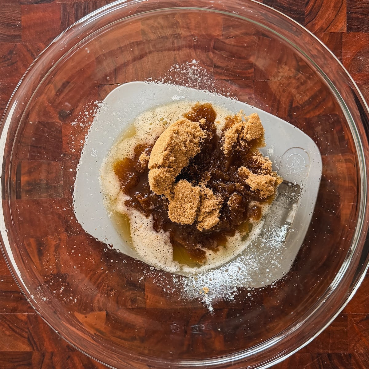 A glass mixing bowl on a wooden surface contains melted butter and packed brown sugar, ready to be mixed together.