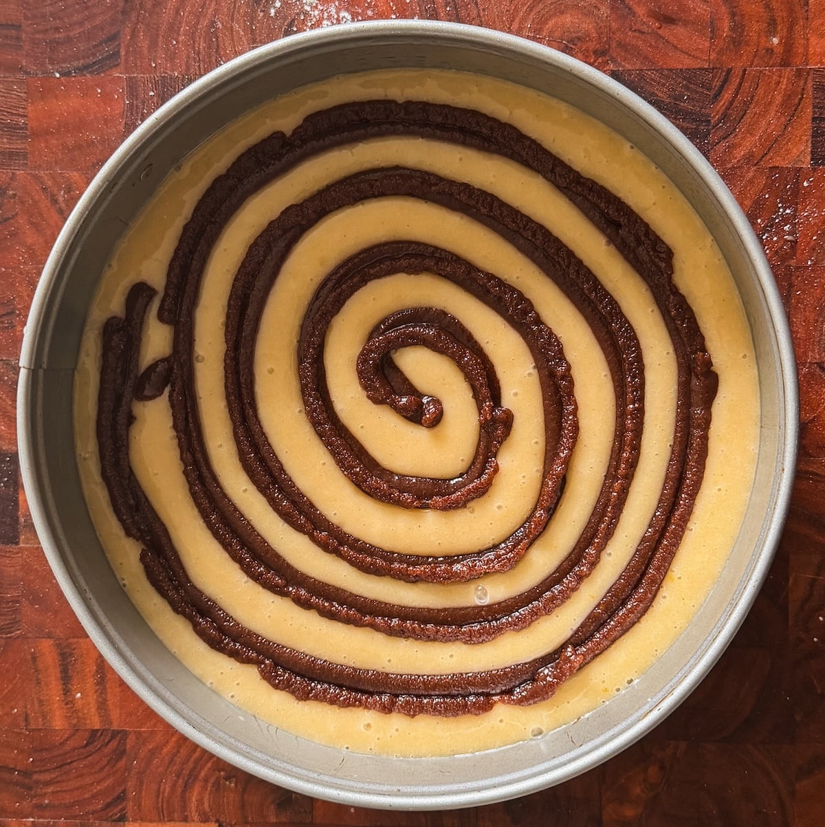 A round cake pan filled with light-colored batter, topped with a dark brown cinnamon sugar spiral swirl, sitting on a wooden surface.