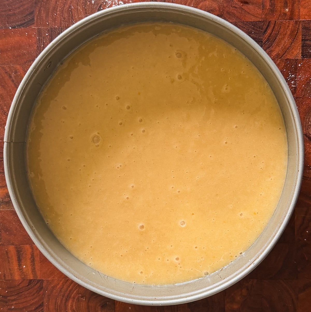A round metal cake pan filled with smooth, light yellow cake batter sits on a dark wooden surface. The batter appears evenly spread and ready for baking.