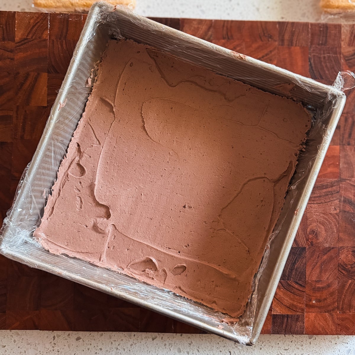 A square metal pan lined with plastic wrap and filled with smooth chocolate ice cream sits on a wooden cutting board.