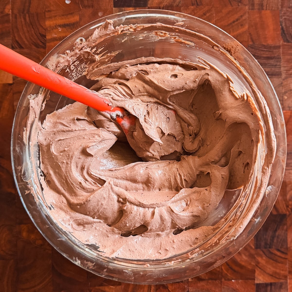 A glass bowl filled with creamy chocolate ice cream being mixed with a red spatula, set on a wooden surface.