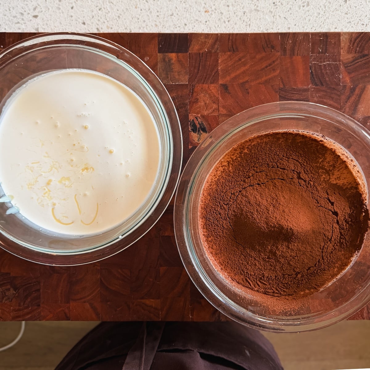 Two glass bowls on a wooden countertop: one with whipped cream, the other with sifted cocoa powder. The edge of a persons dark apron is visible at the bottom of the image.