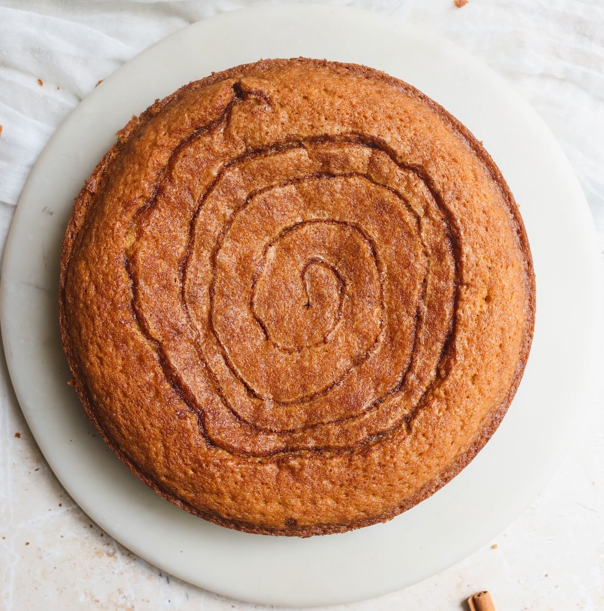 A round, golden-brown cake on a white plate, with a cinnamo sugar swirl in it. The background is a light-colored surface with a white cloth partially visible.