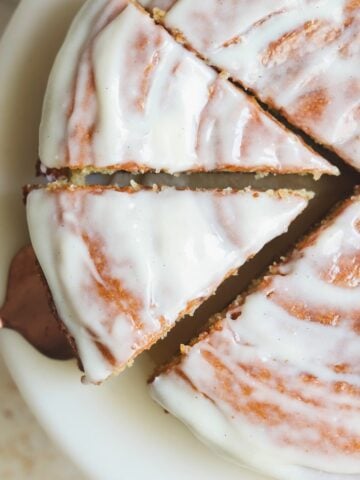 A close-up of a round cinnamon roll cake with white icing, sliced into portions on a white plate. One slice is being lifted with a cake server. The icing has a smooth, glossy finish with visible swirl patterns.