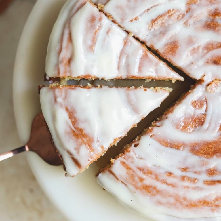 A close-up of a round cinnamon roll cake with white icing, sliced into portions on a white plate. One slice is being lifted with a cake server. The icing has a smooth, glossy finish with visible swirl patterns.