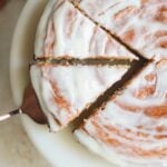 A round cake with white icing, partially sliced, on a white plate. A cake server is lifting one triangular slice, showing the moist texture and smooth glaze on top.