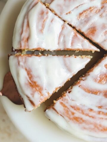 A round cake with white icing, partially sliced, on a white plate. A cake server is lifting one triangular slice, showing the moist texture and smooth glaze on top.