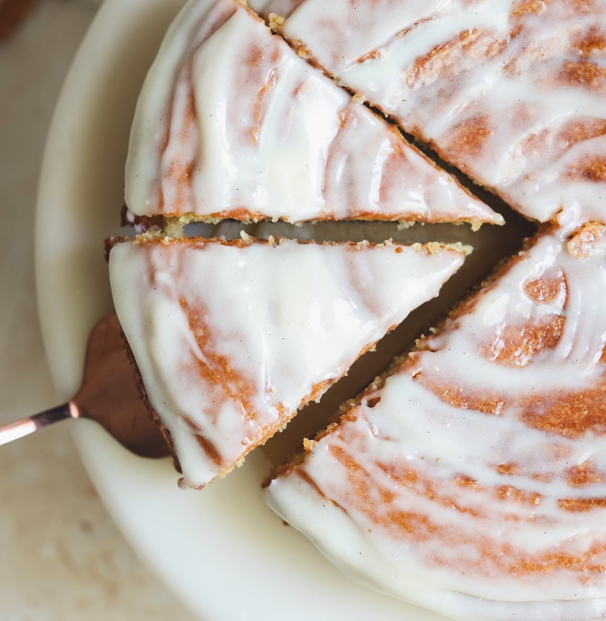 A round cake with white icing, partially sliced, on a white plate. A cake server is lifting one triangular slice, showing the moist texture and smooth glaze on top.