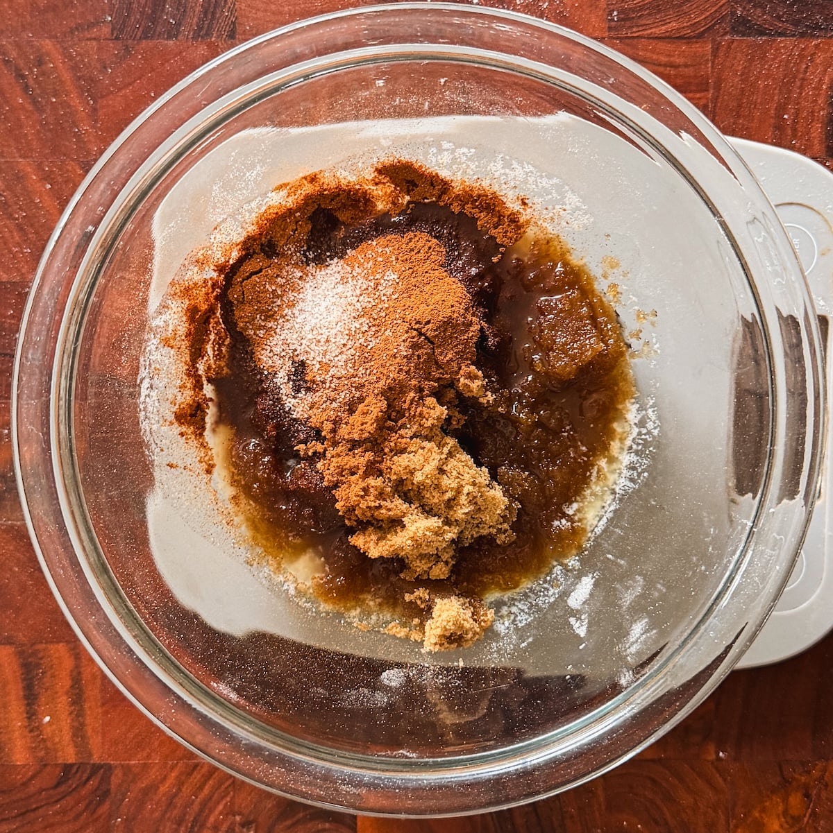 A glass bowl on a wooden surface holds a mixture of brown sugar, ground cinnamon, and melted butter, ready to be mixed for baking.