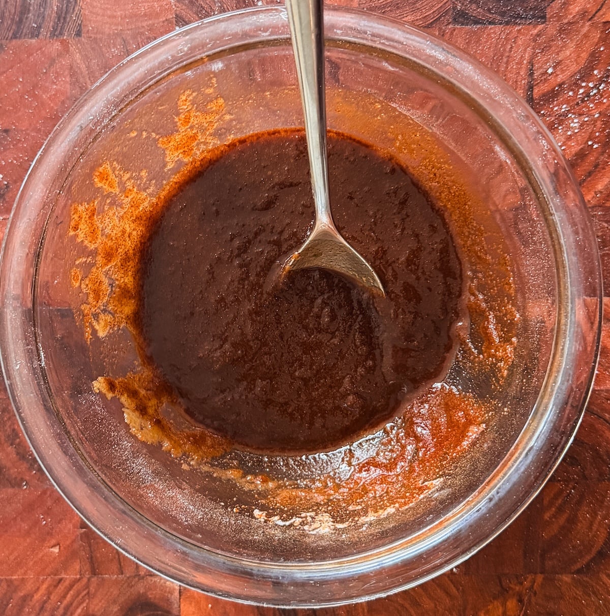 A glass bowl filled with a thick, dark brown mixture sits on a wooden surface. A metal spoon is partially submerged in the mixture, which appears to be in the process of being stirred.