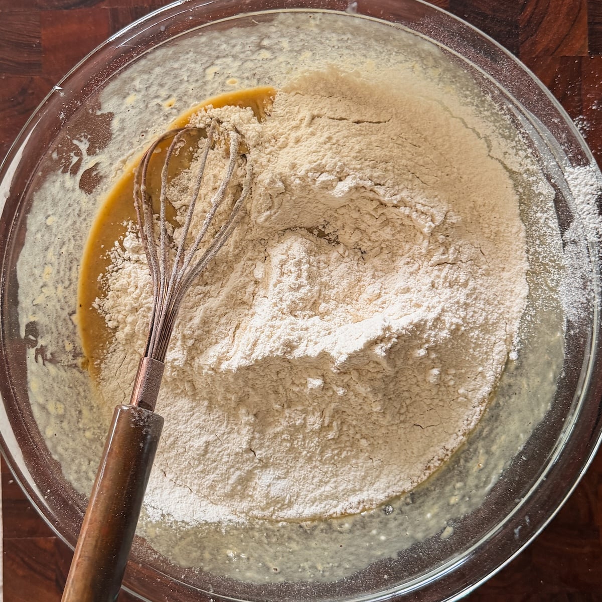 A glass bowl containing flour on top of a wet batter mixture, with a metal whisk resting inside, sits on a wooden surface.
