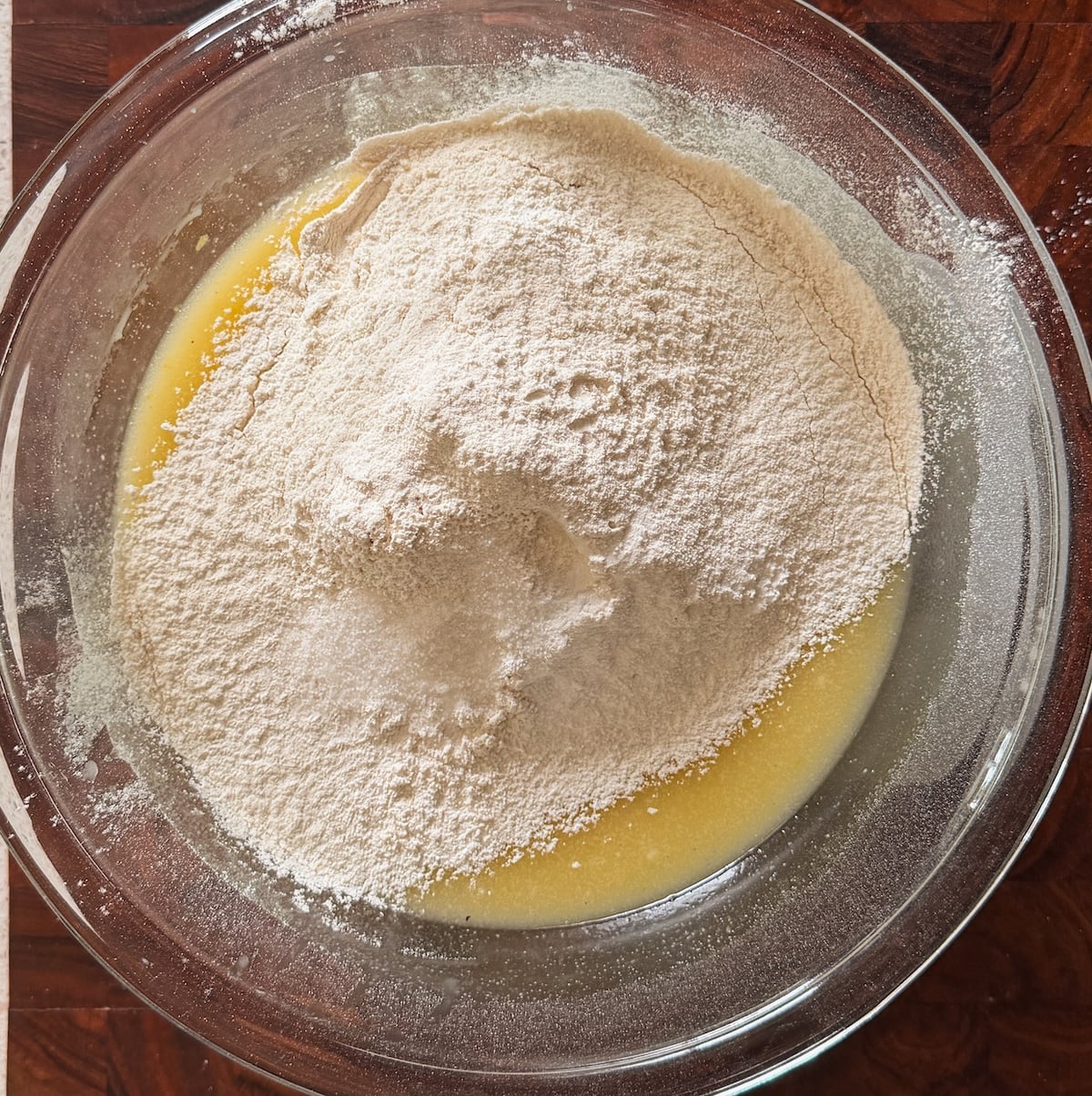 A glass bowl containing a mixture of flour and liquid ingredients, likely for baking, sits on a wooden surface. The flour is piled on top of the wet mixture, not yet stirred in.