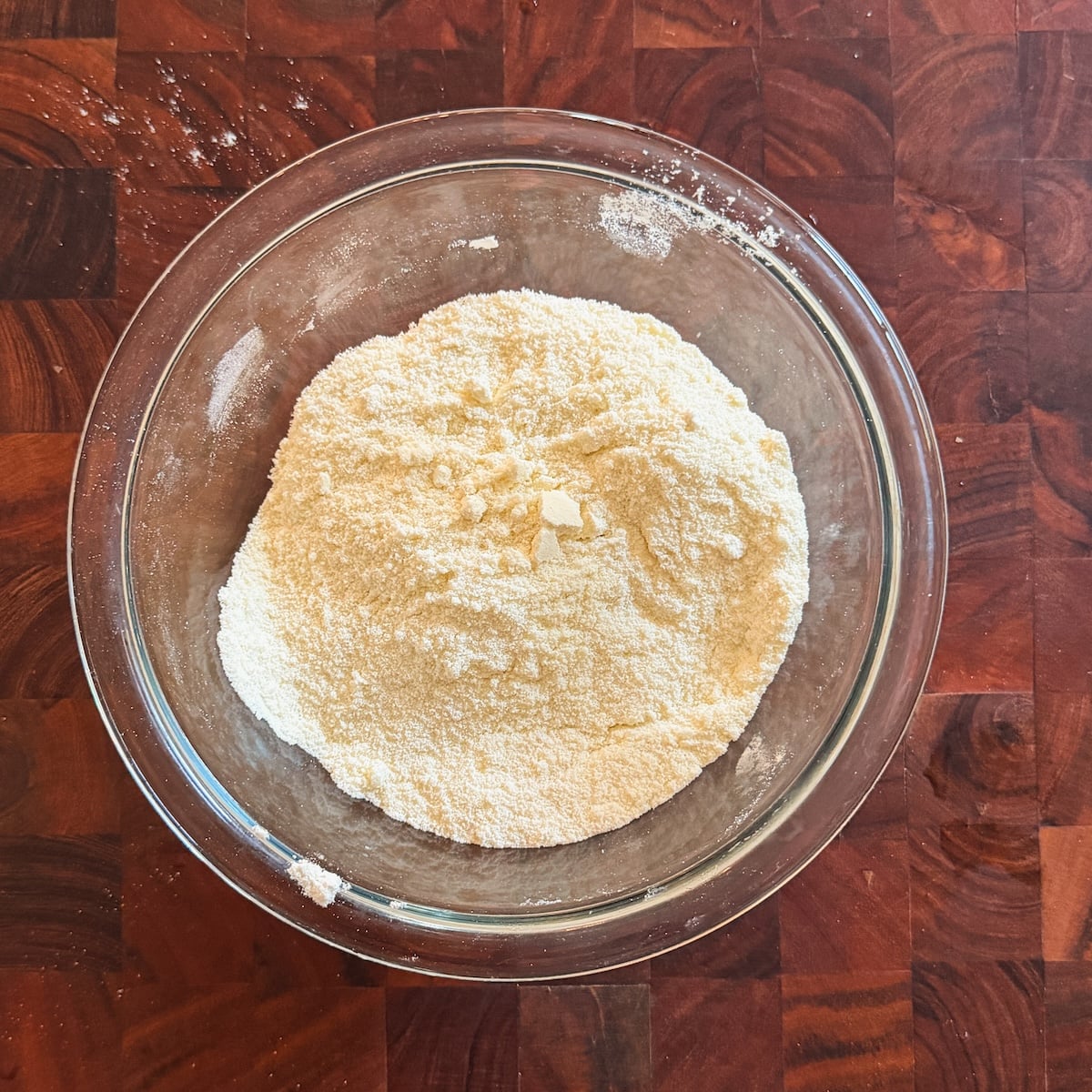 A glass bowl filled with a mound of dry ingredients for gulab jamun sits on a wooden countertop.