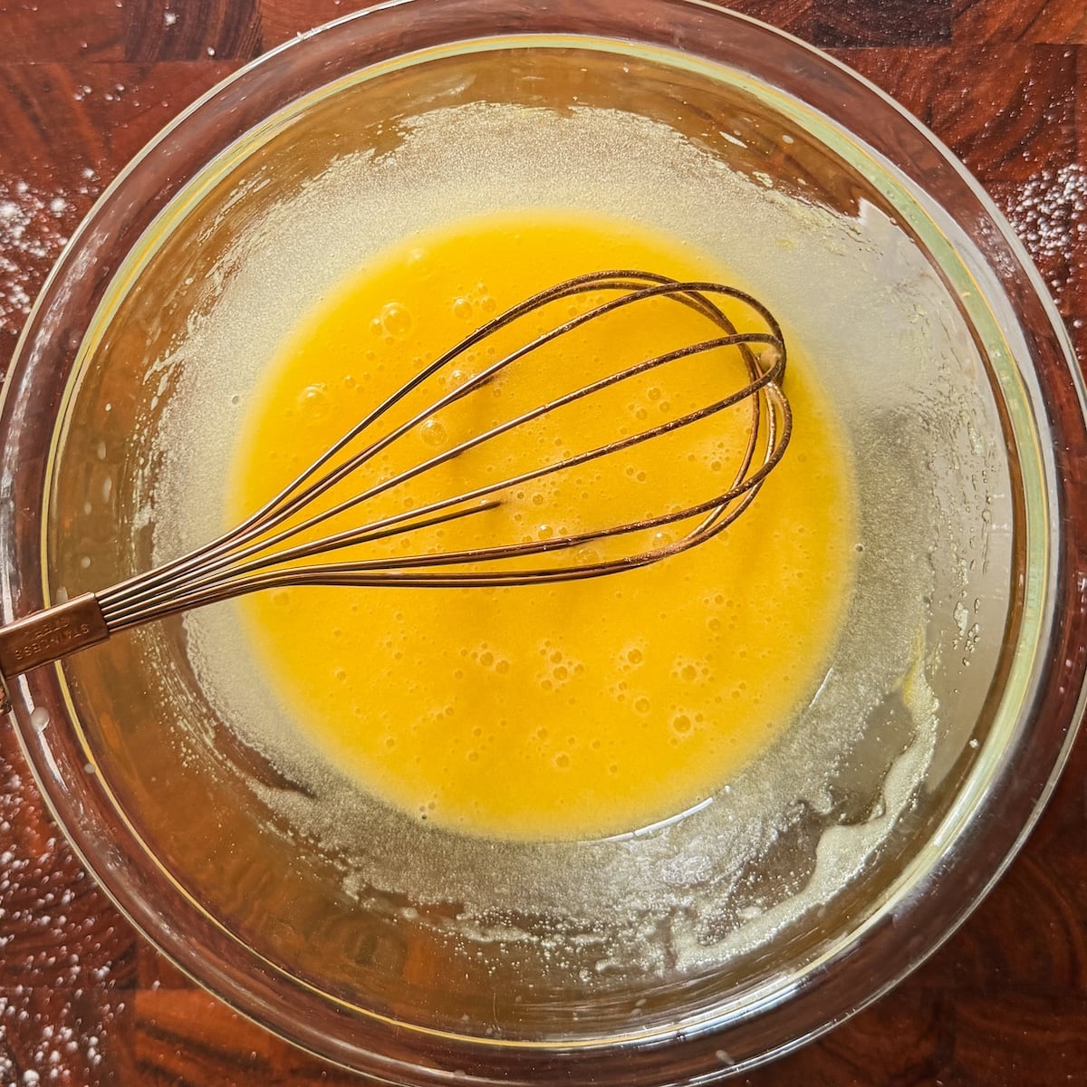 A metal whisk rests in a glass bowl containing a yellow liquid mixture, likely beaten eggs, on a wooden surface lightly dusted with flour.