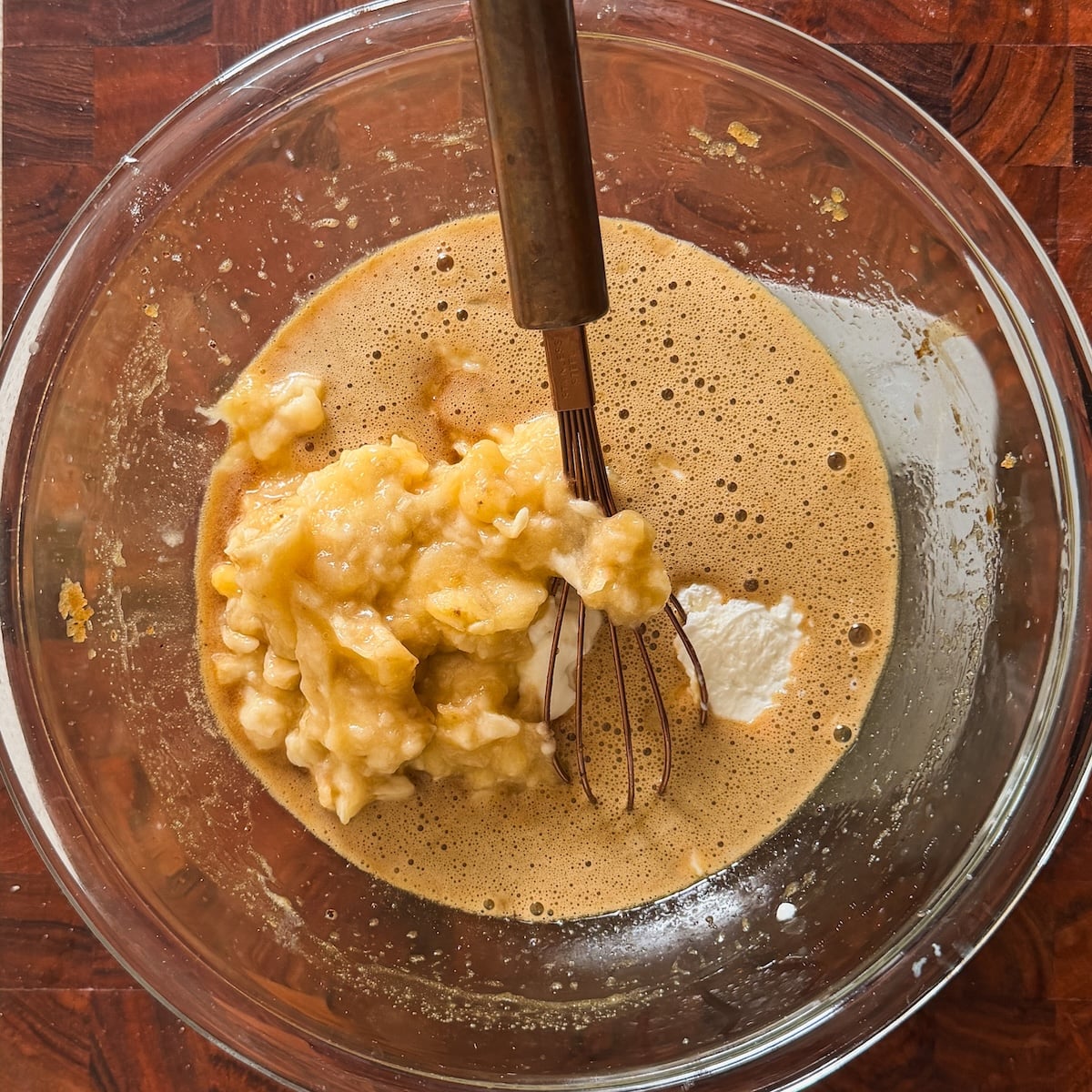 A glass bowl on a wooden surface contains a mixture of mashed bananas, beaten eggs, and a whisk, with some white yogurt visible.