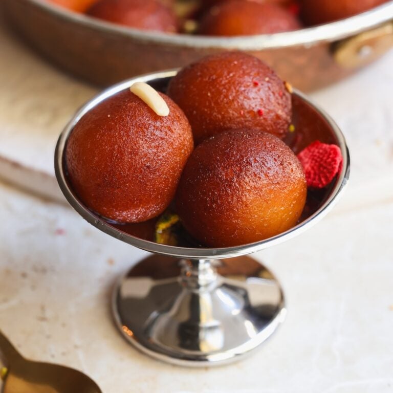 Three round gulab jamuns in a silver dessert bowl, with one topped by a sliver of nut, and a blurred pan of more sweets in the background.