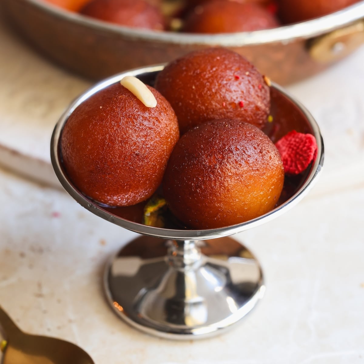 Three round gulab jamuns in a silver dessert bowl, with one topped by a sliver of nut, and a blurred pan of more sweets in the background.
