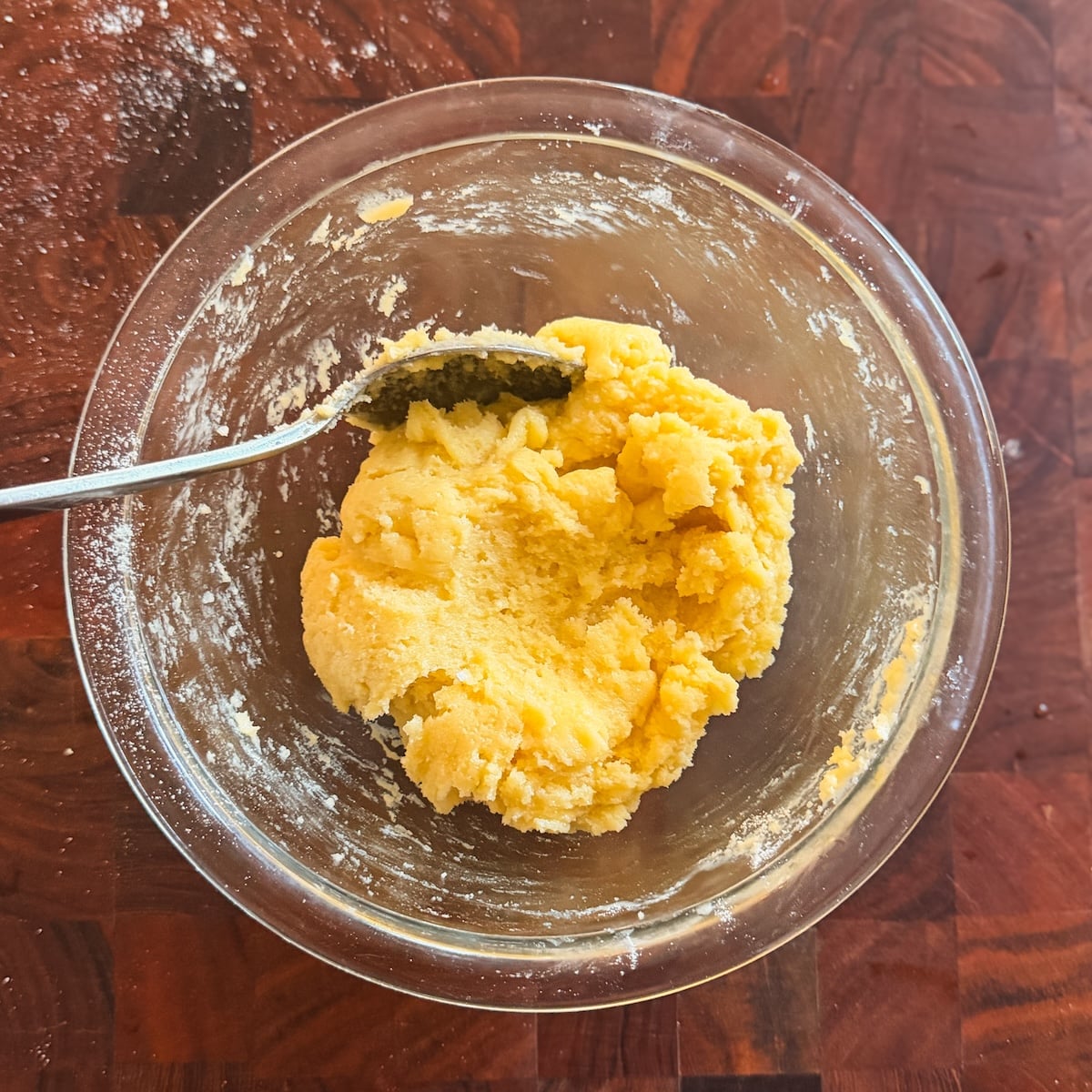 A glass bowl with yellow gulab jamun dough being mixed by a metal spoon, sitting on a wooden surface lightly dusted with flour.