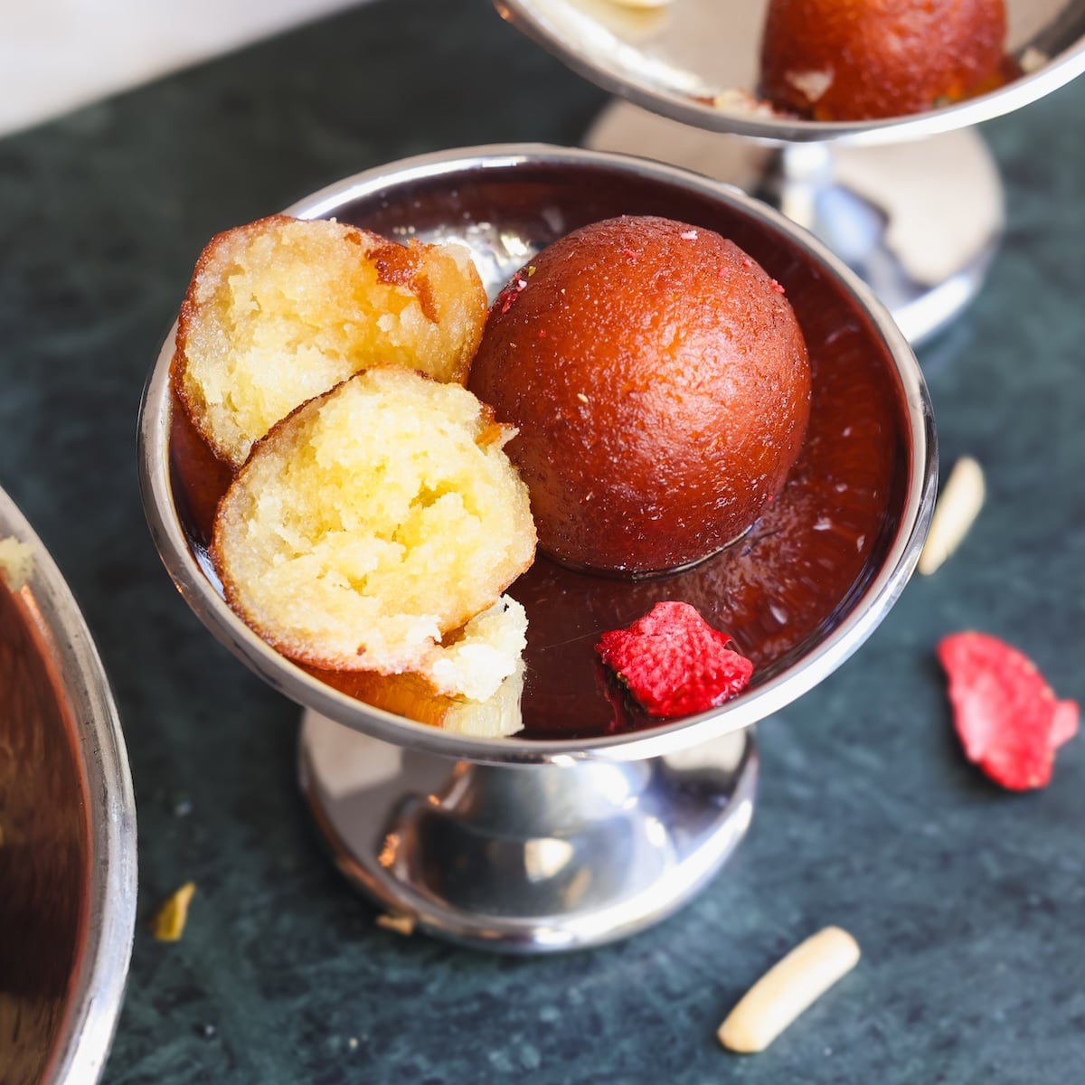 A silver bowl holds one whole and one halved gulab jamun, an Indian dessert, with pieces of dried fruit and nuts on a dark green surface.