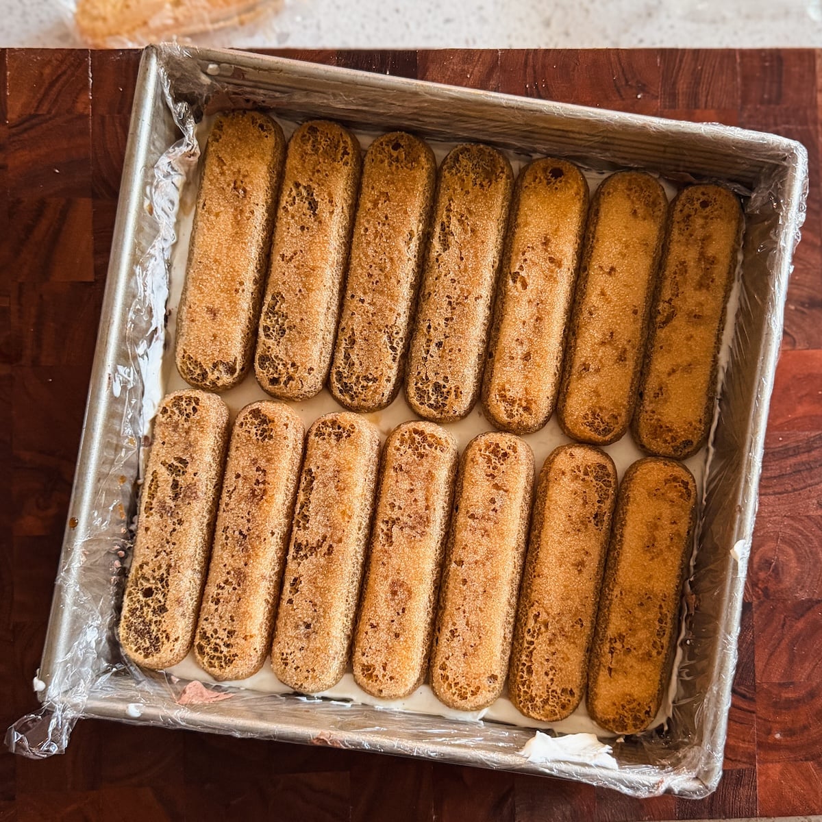 A square baking pan lined with plastic wrap is filled with a layer of ladyfinger cookies arranged neatly side by side over a creamy filling, ready for the next step in making tiramisu.