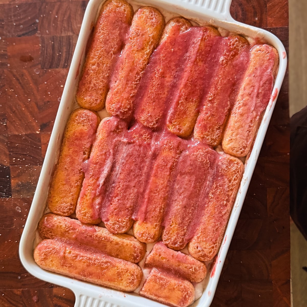 A rectangular white baking dish filled with a single layer of ladyfinger biscuits soaked in a strawberry syrup, arranged neatly on a wooden surface.