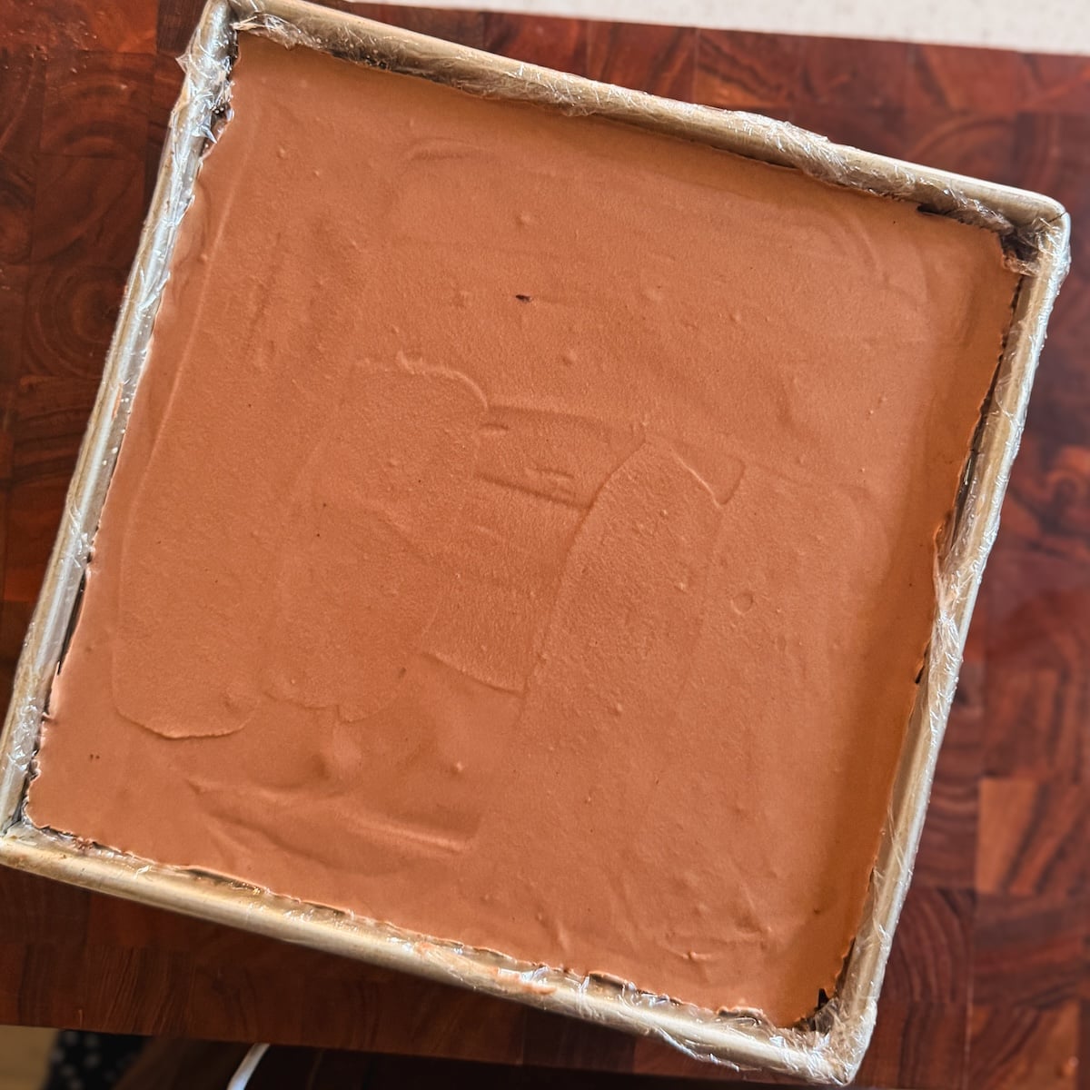 A square pan lined with plastic wrap, filled with smooth chocolate mousse, sits on a wooden surface. The top is evenly spread with visible spatula marks.