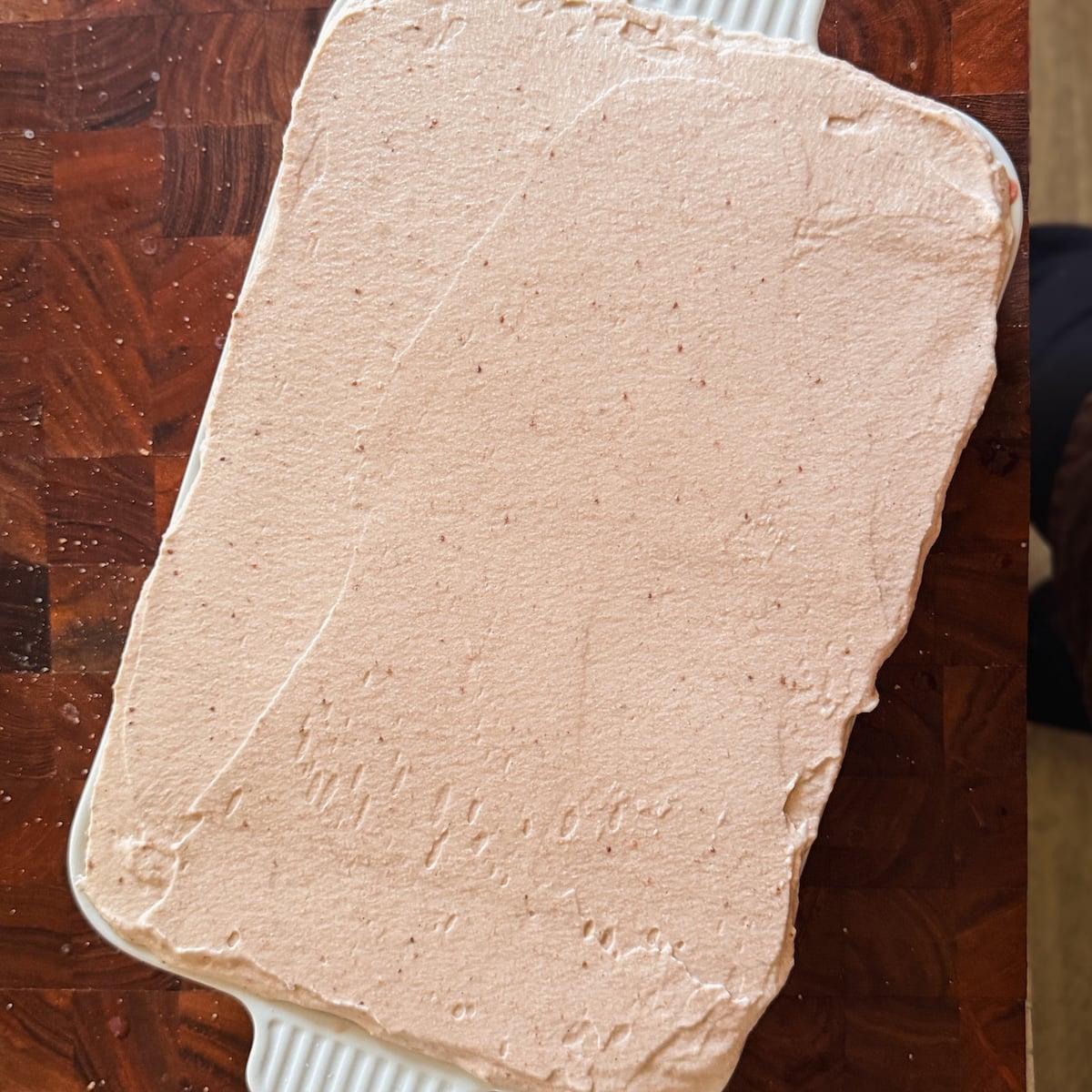 A rectangular dish filled with a dessert topped with a smooth, light pink cream, sitting on a wooden table. The frosting appears evenly spread across the surface.