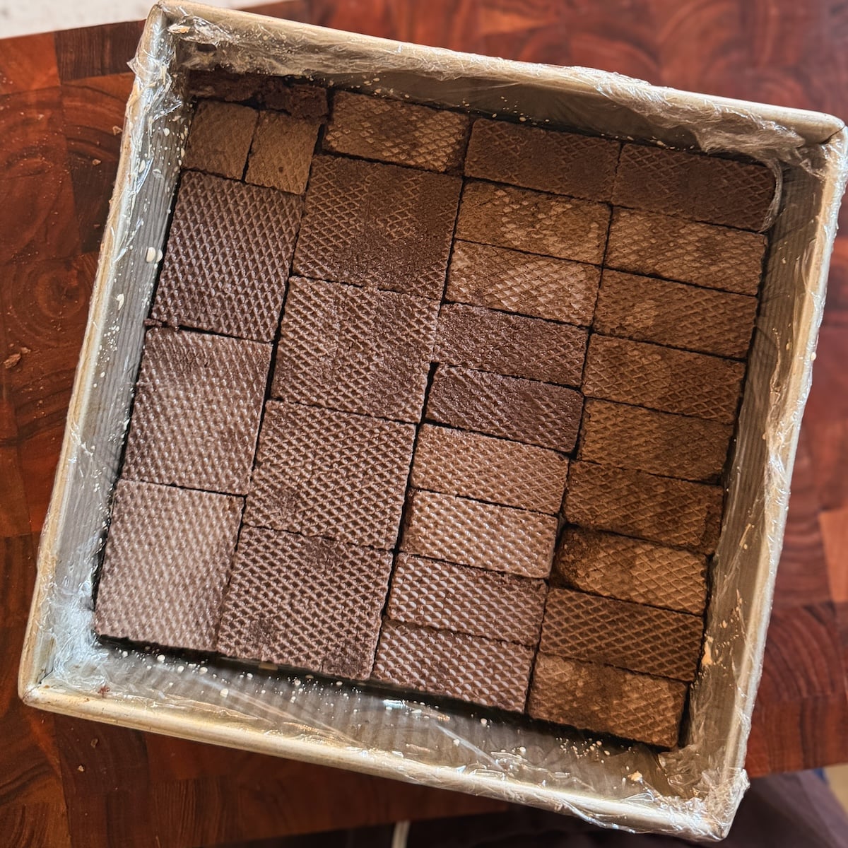 A box lined with clear plastic wrap is filled with neatly arranged rectangular chocolate wafers of varying shades of brown, stacked in a grid pattern.