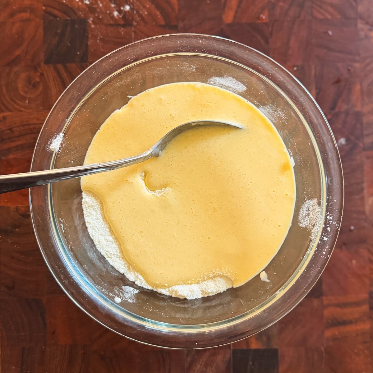 A glass bowl containing a yellow liquid mix being mixed with dry ingredients, with a metal spoon resting inside, sits on a wooden surface.