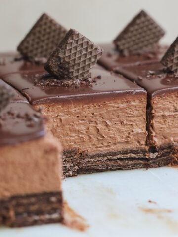 Close-up of sliced chocolate mousse cake with a biscuit base, topped with a glossy chocolate glaze and decorated with pieces of chocolate wafer cookies. One slice is partially removed, revealing the creamy texture.