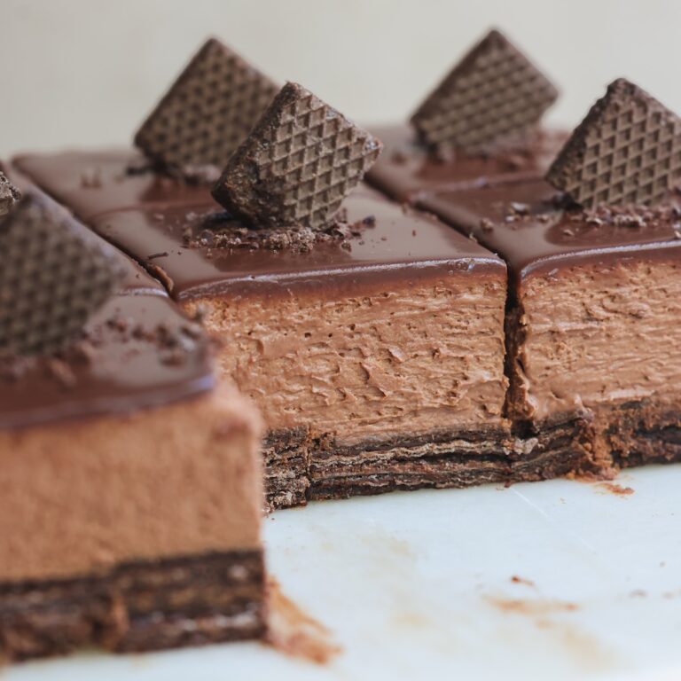 Close-up of sliced chocolate mousse cake with a biscuit base, topped with a glossy chocolate glaze and decorated with pieces of chocolate wafer cookies. One slice is partially removed, revealing the creamy texture.