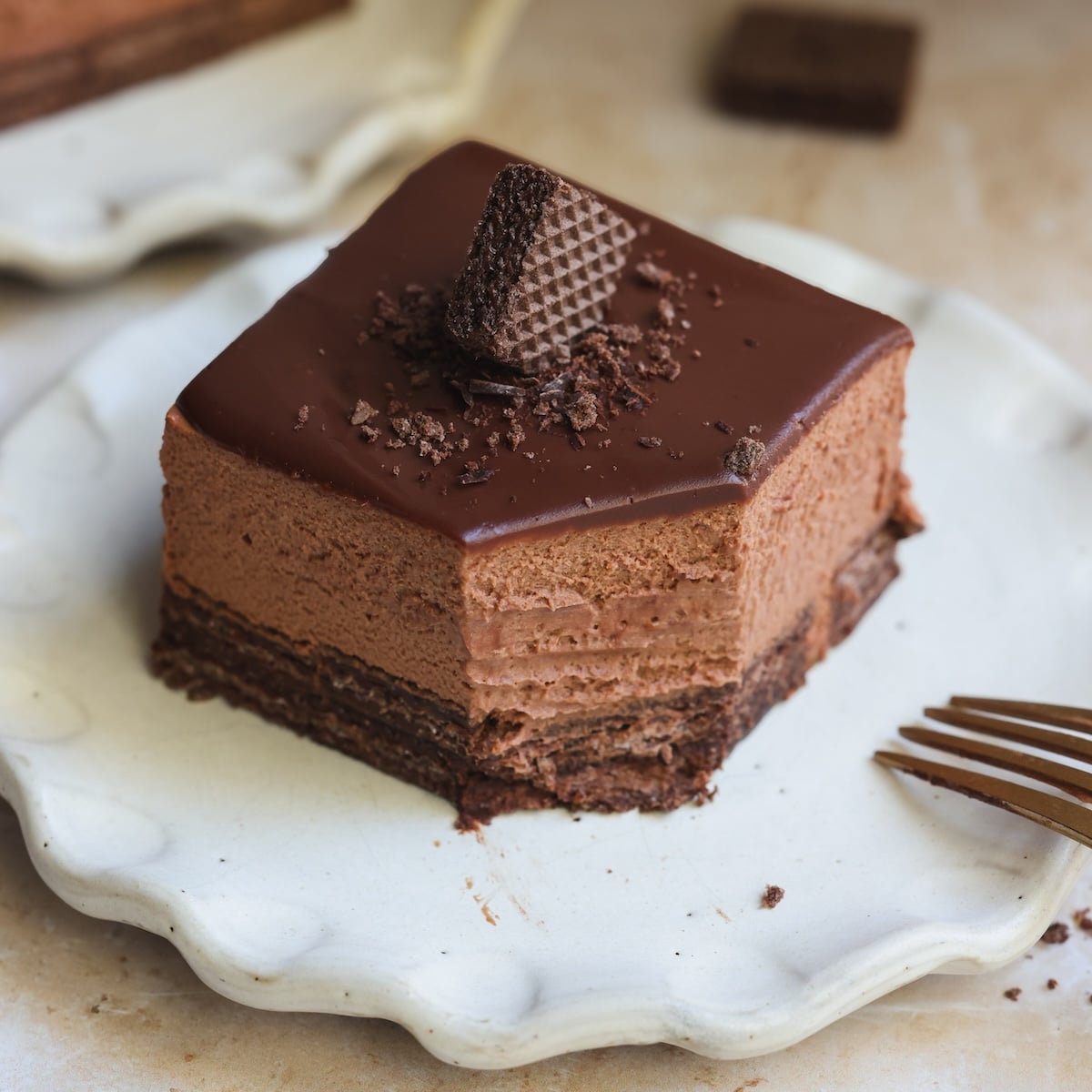 A square slice of no bake chocolate mousse cake with a glossy chocolate ganache, topped with a chocolate wafer and chocolate shavings, served on a white plate beside a gold fork.