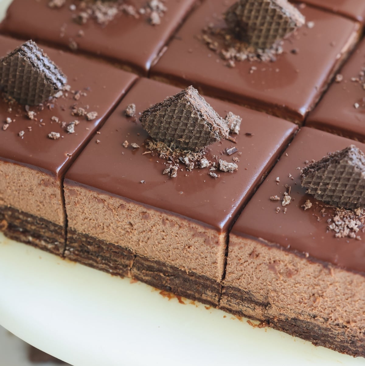 Close-up of sliced chocolate mousse cake squares with a glossy chocolate topping, each piece topped with a small chocolate wafer and chocolate crumbs on a white surface.
