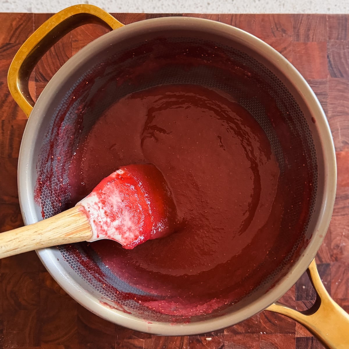 A saucepan with gold handles sits on a wooden surface, containing a thick, dark red mixture being stirred with a red spatula featuring a wooden handle.