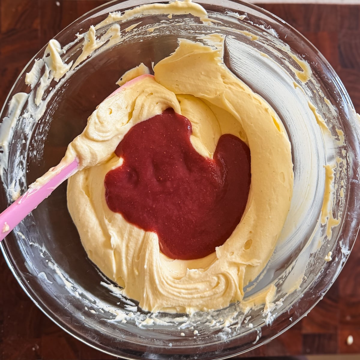 A glass bowl with pale yellow mix and a pink spatula. A portion of red fruit puree is poured on top of the batter, ready to be mixed. The bowl sits on a wooden surface.