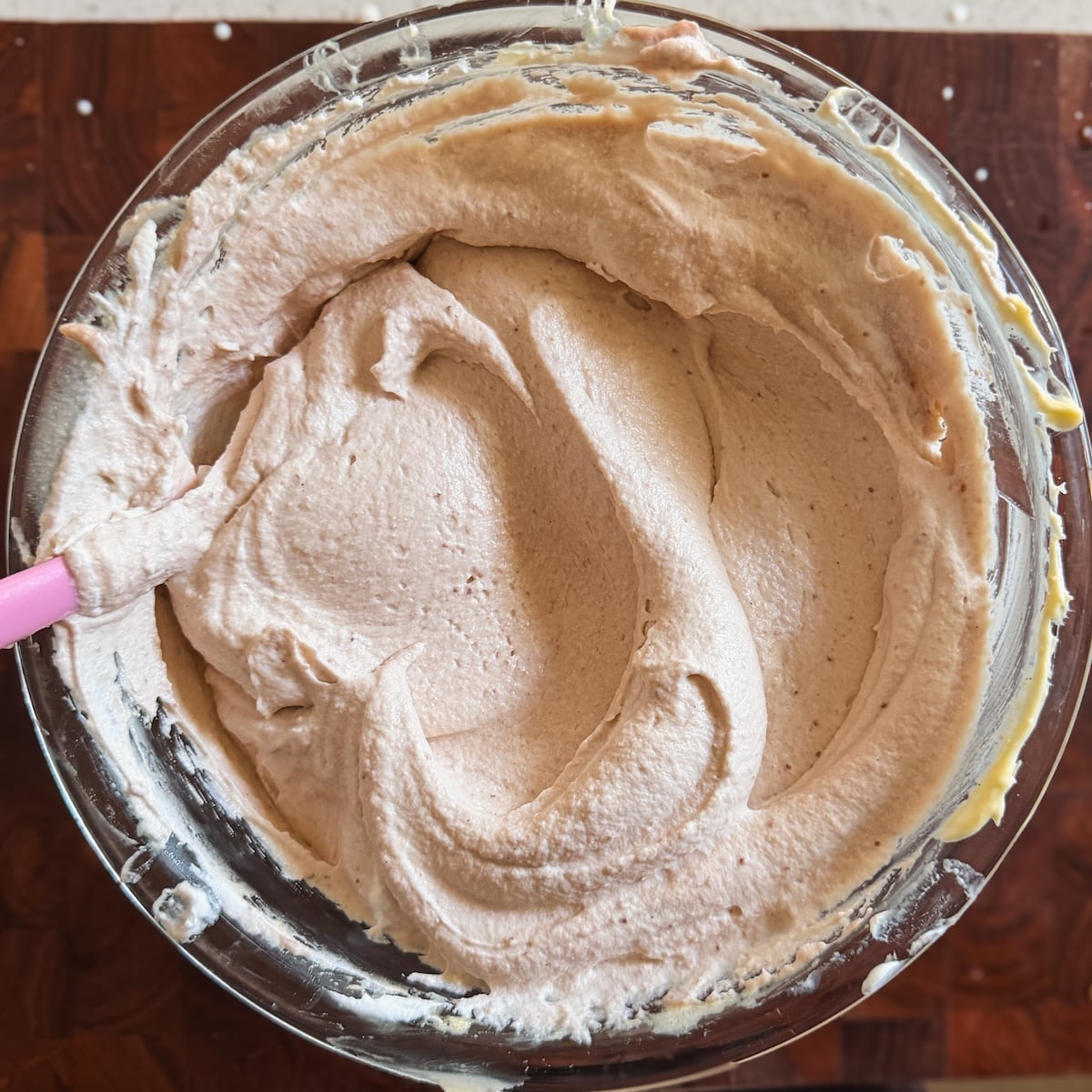A glass bowl filled with light pink whipped frosting or batter sits on a wooden surface, with a pink spatula resting on the side of the bowl.