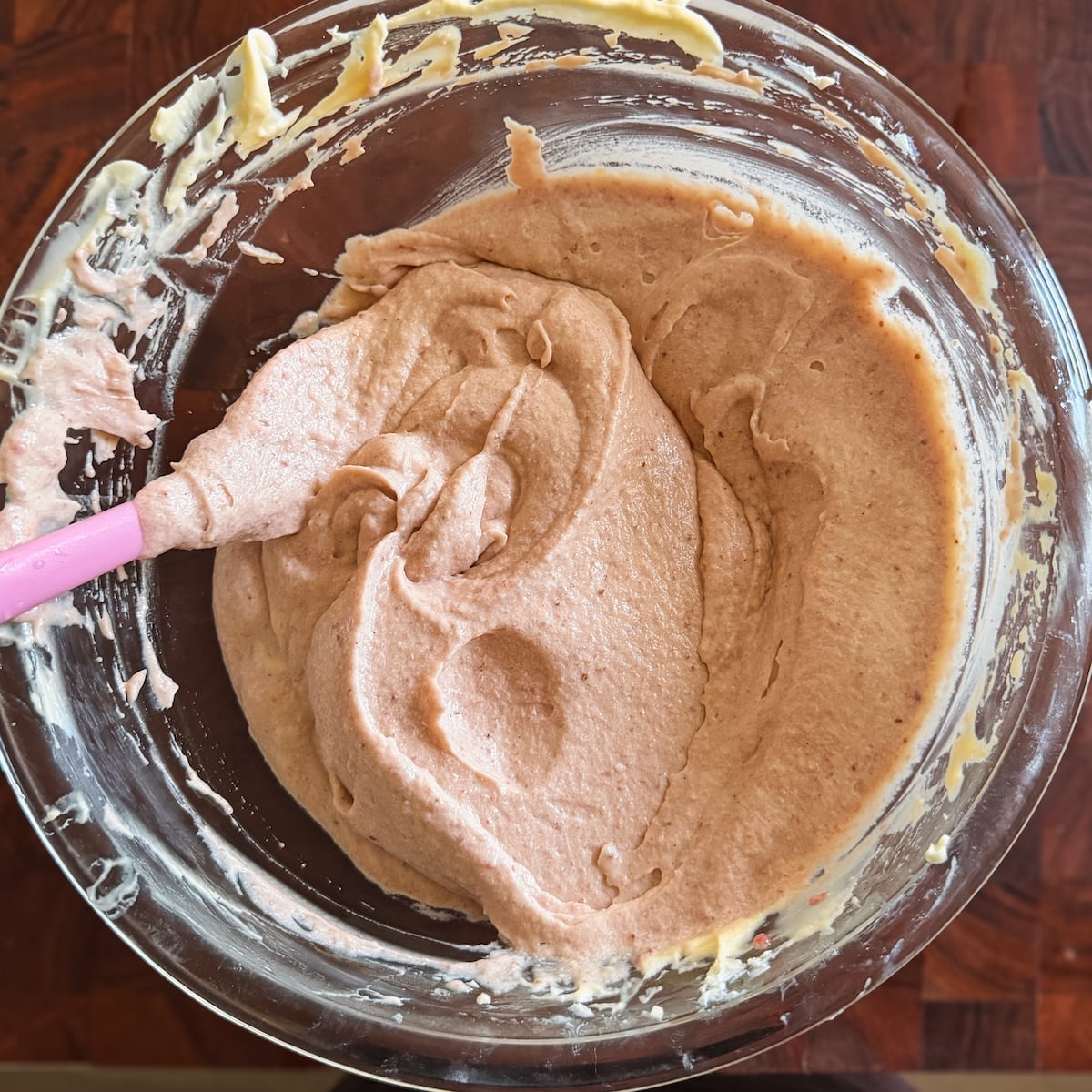 A glass bowl containing light pink mixture being mixed with a pink spatula, set on a wooden surface. The batter appears smooth and creamy, ready for baking.