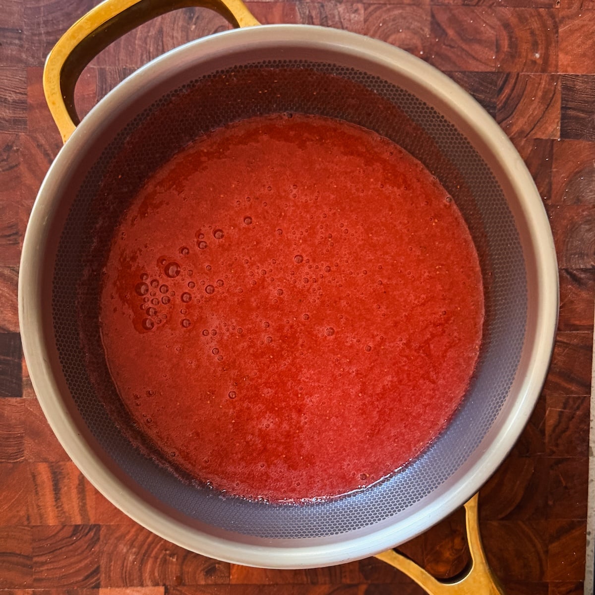 A pot with light-colored handles sits on a wooden surface, filled with a smooth, red liquid that appears to be pureed strawberries