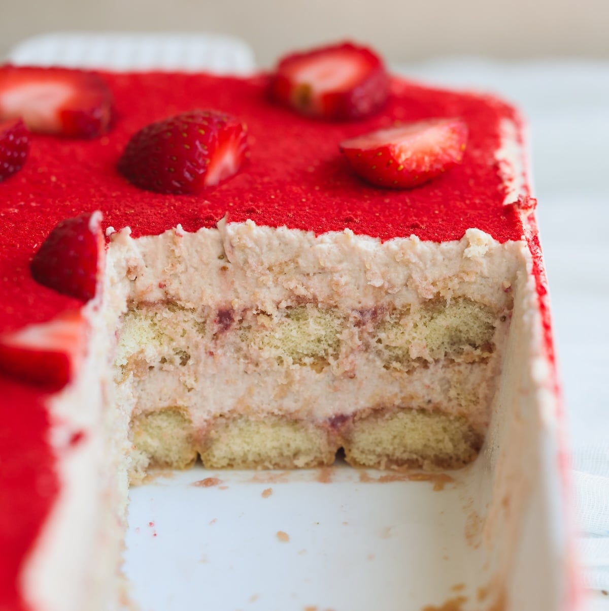 A rectangular strawberry tiramisu with layers of cream and ladyfingers, topped with bright red strawberries. A large slice has been removed, showing the cakes layered interior.