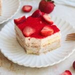 A slice of strawberry tiramisu topped with fresh strawberry halves sits on a white scalloped plate, with a whole strawberry and another dessert piece in the background.