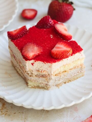 A slice of strawberry tiramisu topped with fresh strawberry halves sits on a white scalloped plate, with a whole strawberry and another dessert piece in the background.