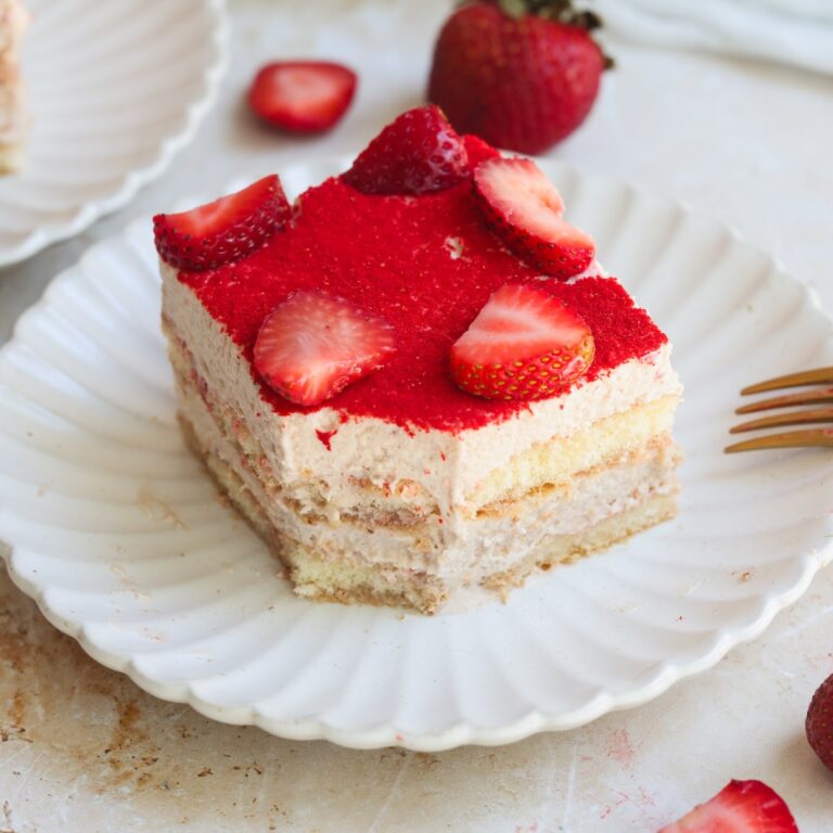A slice of strawberry tiramisu topped with fresh strawberry halves sits on a white scalloped plate, with a whole strawberry and another dessert piece in the background.