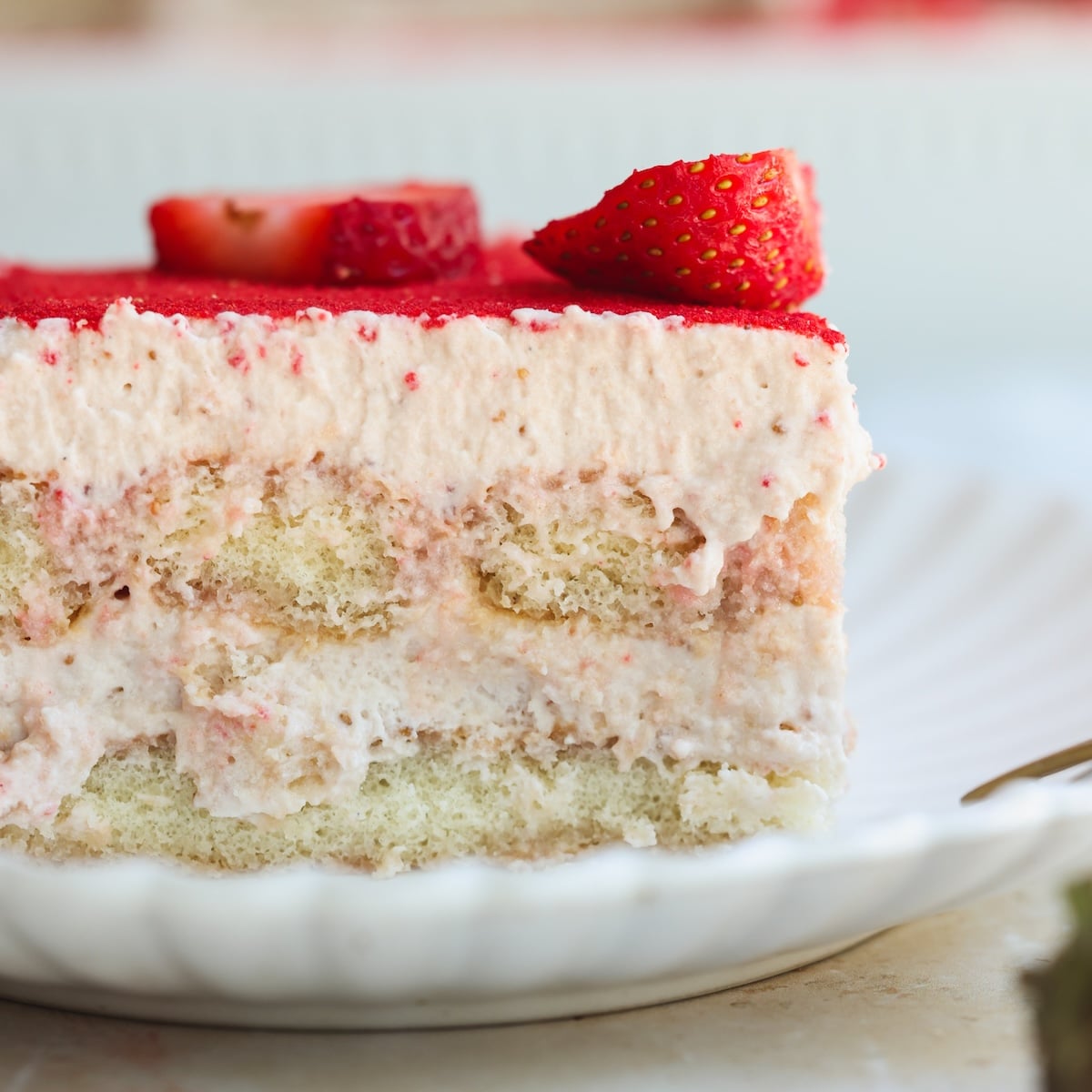 A close-up of a slice of strawberry tiramisu on a white plate, showing layers of creamy filling, sponge cake, and strawberry topping, garnished with fresh strawberry pieces.