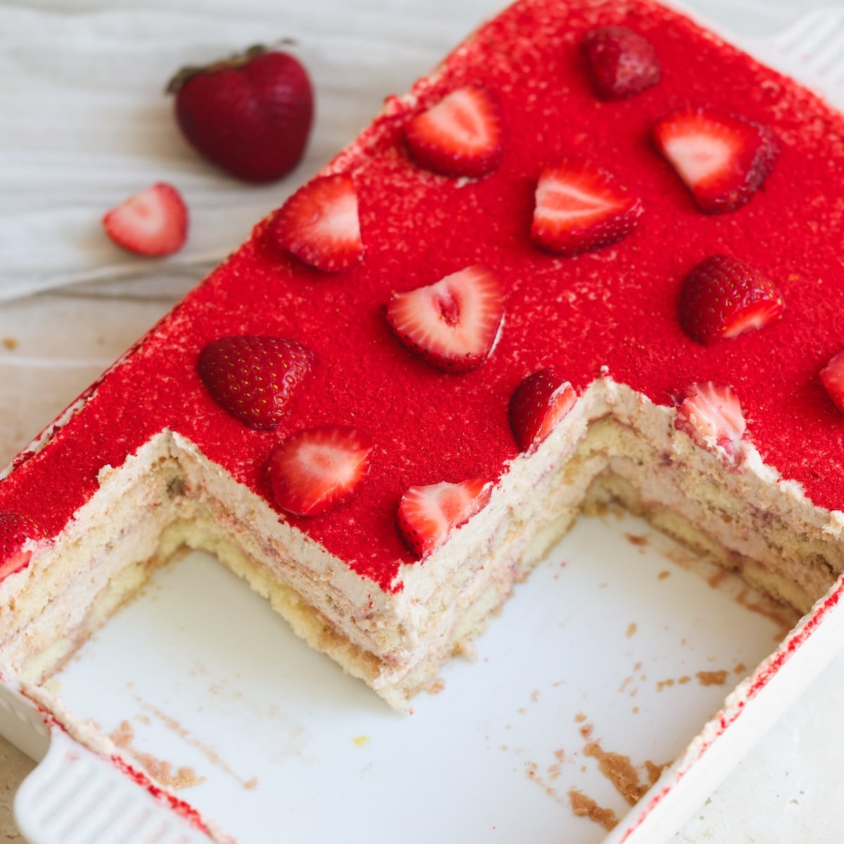 A rectangular dish of strawberry tiramisu with a large square piece missing, showing layers of cream and cake. The top is covered in vibrant red strawberry powder and sliced strawberries. Two whole strawberries are nearby.