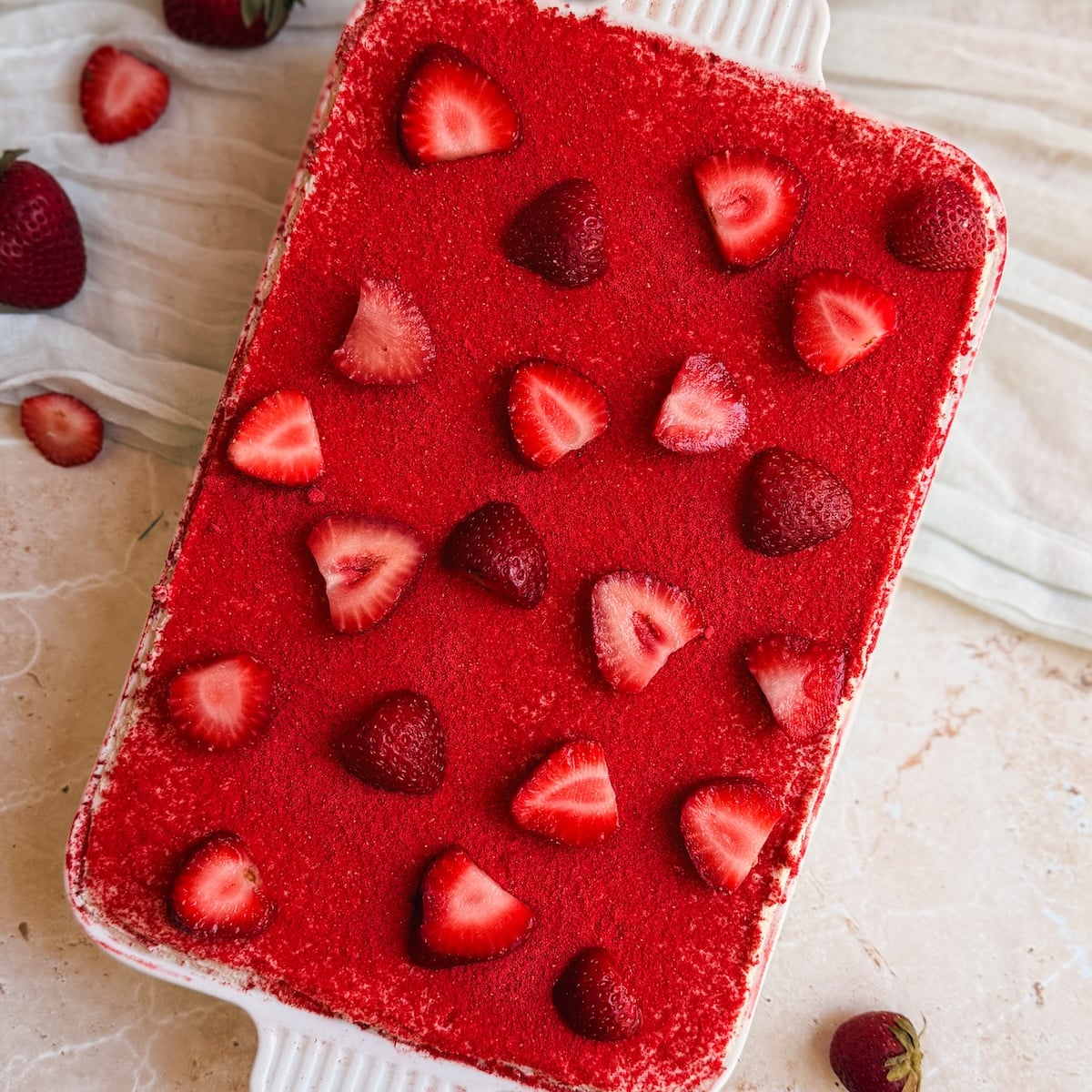 A rectangular dish filled with a bright red dessert, topped with whole and sliced strawberries, sits on a light surface next to a cloth and a few loose strawberries.
