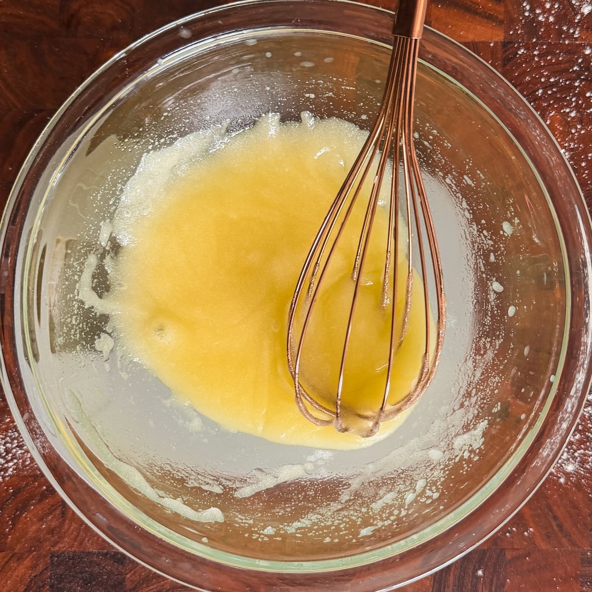 A glass bowl containing a yellow mix being whisked with a copper whisk, set on a wooden surface with some flour sprinkled around.
